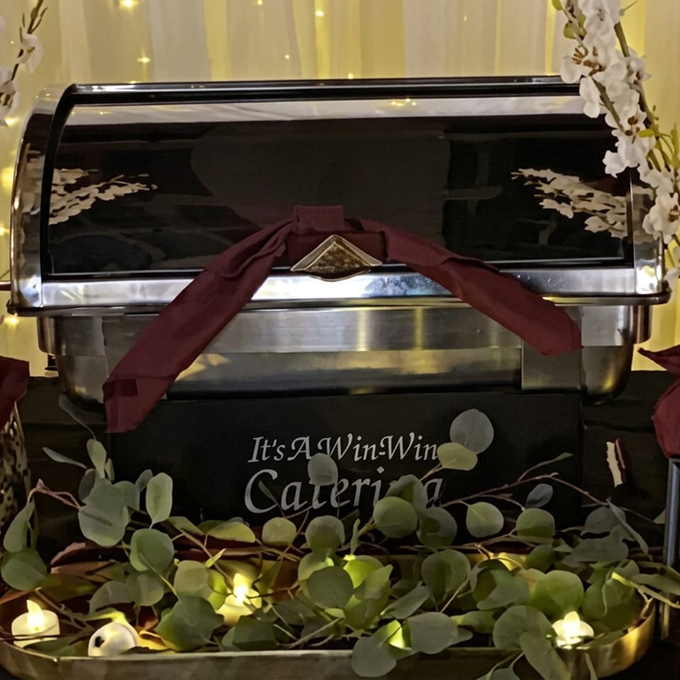 Decorated catering buffet station with a stainless chafing dish, burgundy napkins, white flowers, greenery, candles, and a grilled beef tenderloin sign.