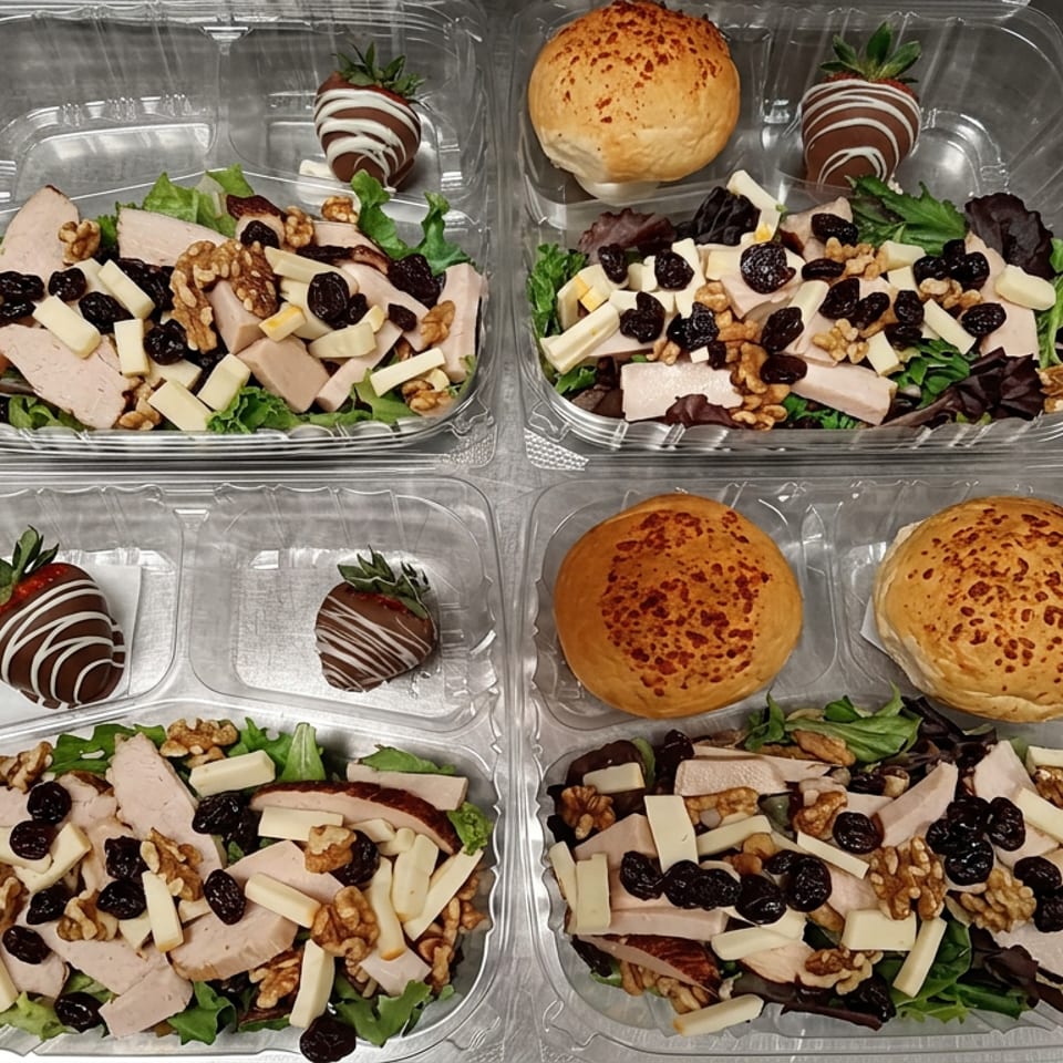 Four takeout containers hold leafy salads with sliced turkey, cheese, walnuts, and dried cranberries, alongside rolls and chocolate-covered strawberries.
