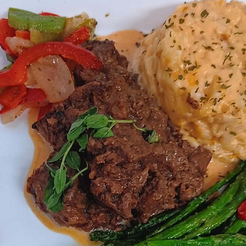 Plated sliced beef with creamy herb-seasoned rice, sautéed peppers and onions, and green vegetables on a white dish.