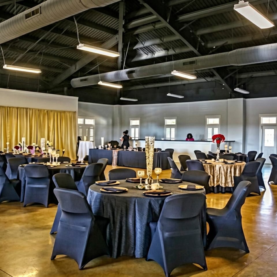 Elegant event hall with round tables, navy linens, gold centerpieces, and a draped golden stage backdrop.