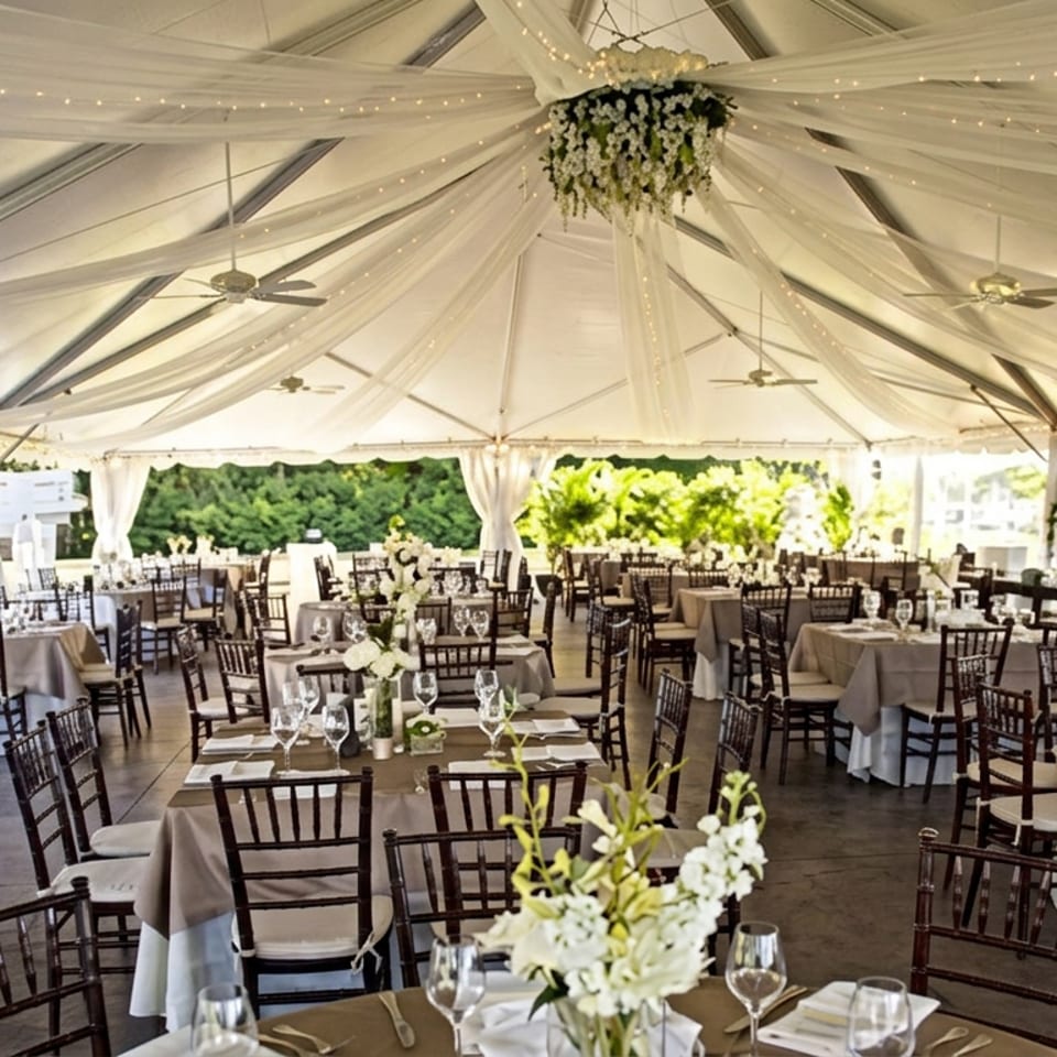 Elegant outdoor wedding tent with draped fabric, string lights, flower arrangements, and round tables set for dining.