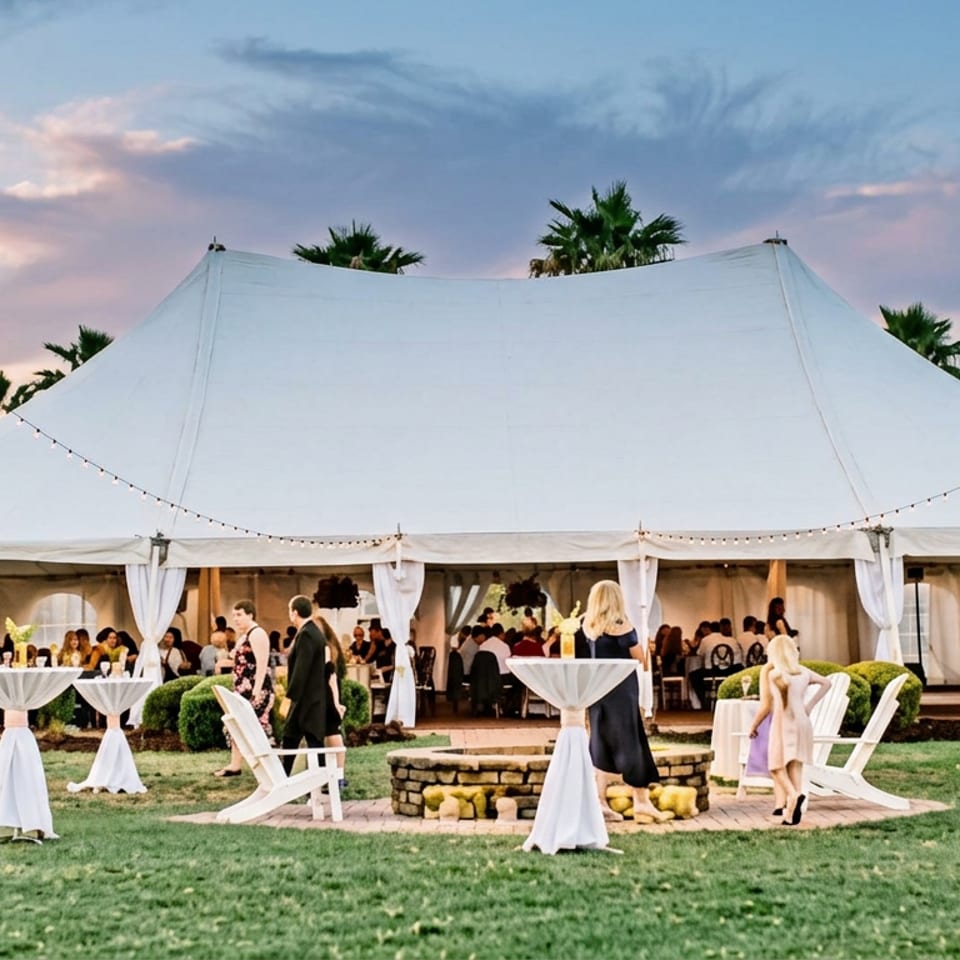 Elegant outdoor wedding reception under a large white tent, with guests, cocktail tables, palm trees, and string lights at sunset.