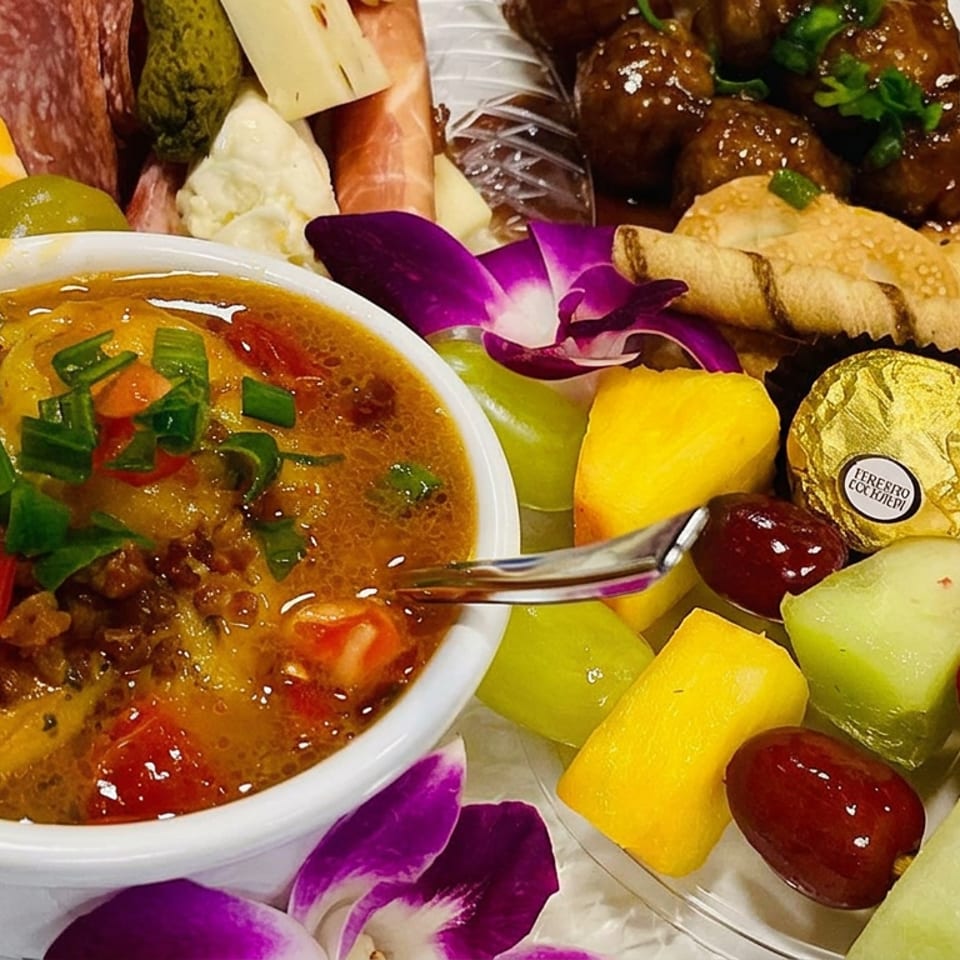 Appetizer platter featuring a savory dip, charcuterie, meatballs, spring rolls, crackers, fresh fruit, chocolate, and orchid garnish.
